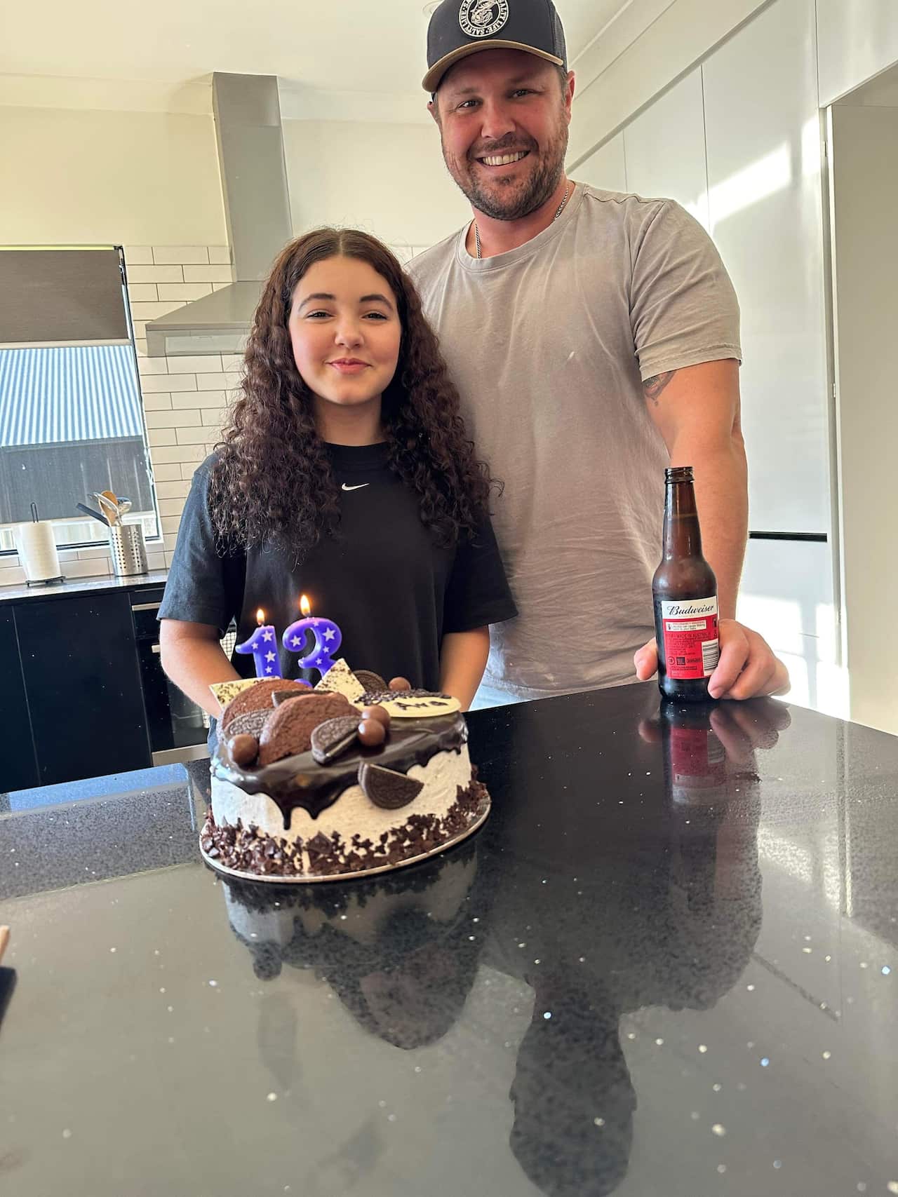 A man standing in a kitchen with his teen daughter beside him and a birthday cake on the bench in front of them.